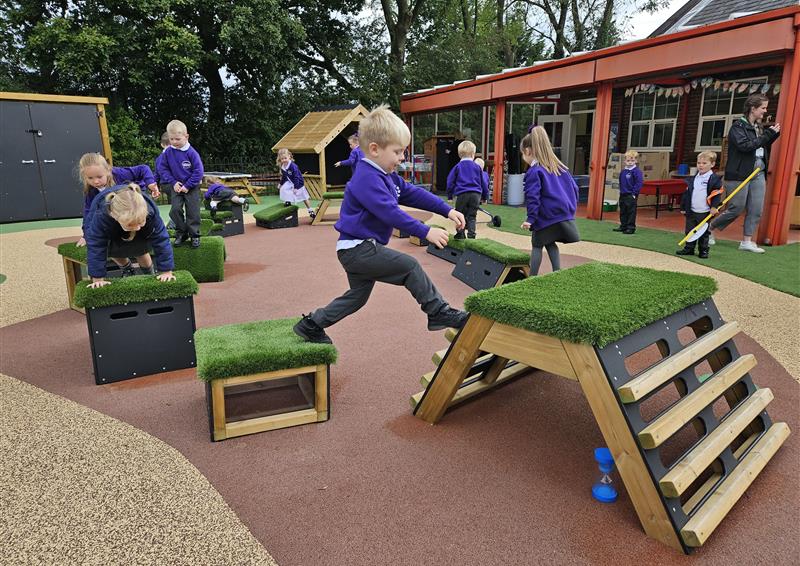 Wooden blocks with artificial grass tops are placed in an oval. Each block is a different shape. 10 children are jumping between each block as they move in anti-clockwise matter.